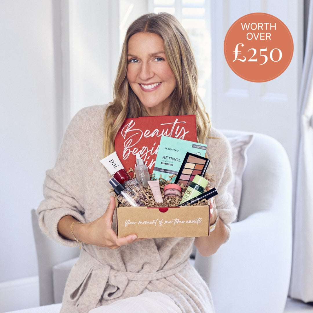 A smiling woman in a beige robe holds a cardboard gift box filled with assorted beauty products, including a red 'Beauty begins' card, makeup palettes, and skincare items. She is seated in a bright white chair in a softly lit room.