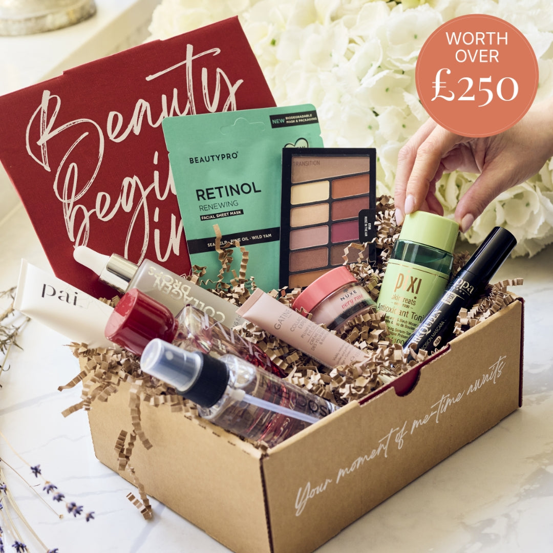 A curated beauty gift box filled with multiple cosmetic products, including a red Beauty Begins set folder, a green Retinol Cleansing sheet mask, a makeup palette, a green P.I XI bottle, and other tubes and bottles, arranged in brown paper shred inside a cardboard box.