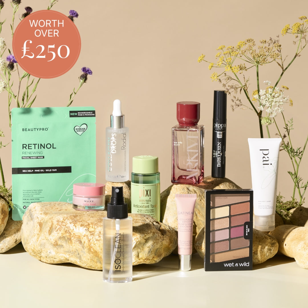 A curated beauty product arrangement on a beige surface with decorative rocks and flowers. Visible products include a mint green Beautypro Retinol sheet mask, a clear dropper bottle labeled KVD, a pink jar, a green bottle labeled KIXI, a black mascara tube labeled KVD Beauty, a white tube labeled pat, a clear bottle labeled SOELOS, a pink tube, a black-and-white wet n wild eyeshadow palette, and a round pink item.